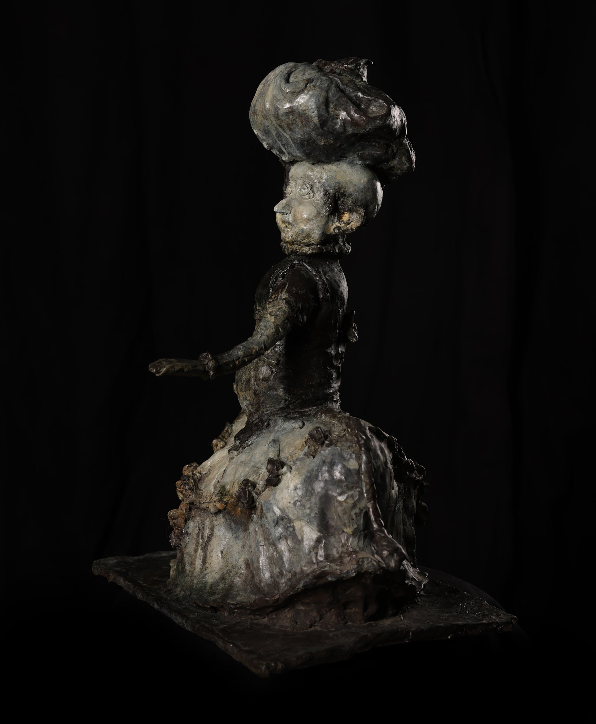 Metallic sculpture of a figure with a large head and detailed dress on a black background