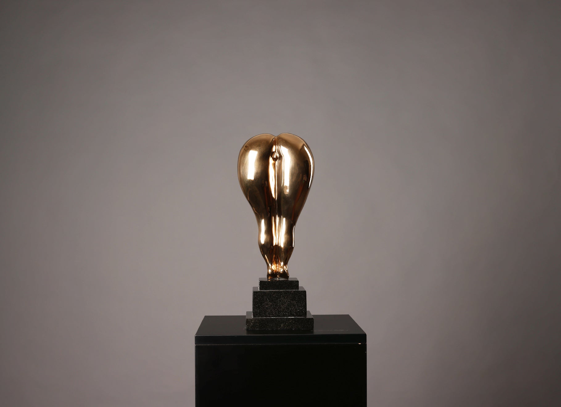 Gold light bulb sculpture on a black pedestal against a gray background