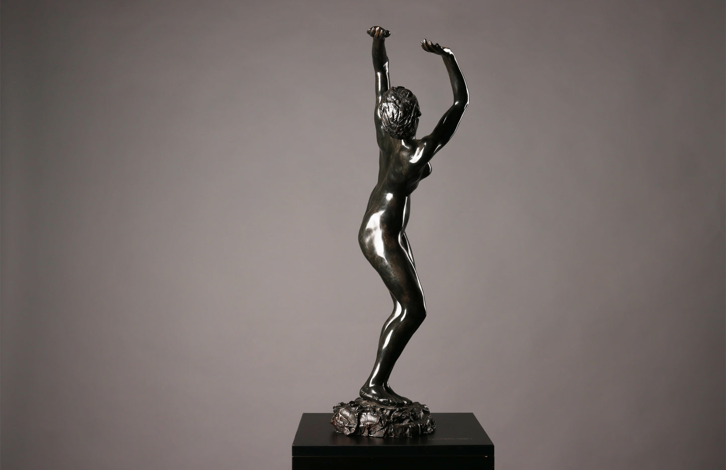 Bronze statue of a nude figure with raised arms on a dark pedestal against a gray background
