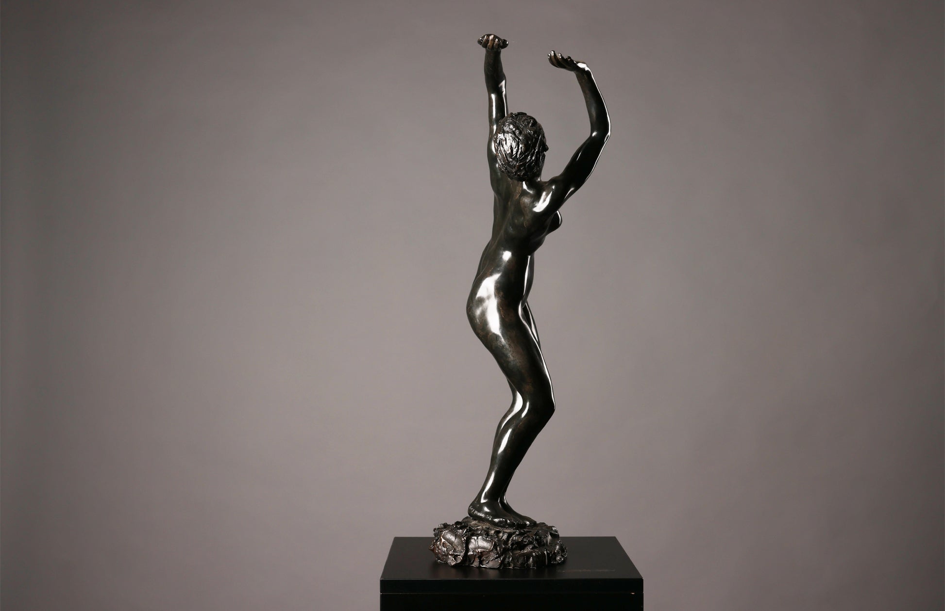Bronze statue of a nude figure with raised arms on a dark pedestal against a gray background