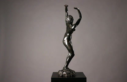 Bronze statue of a nude figure with raised arms on a dark pedestal against a gray background